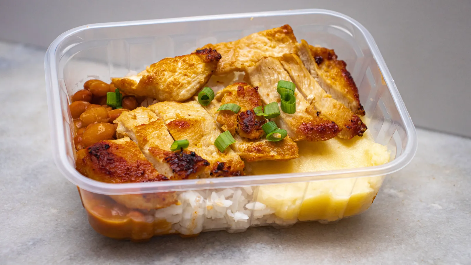 Frango grelhado com arroz, feijão e verduras em um recipiente transparente para viagem.