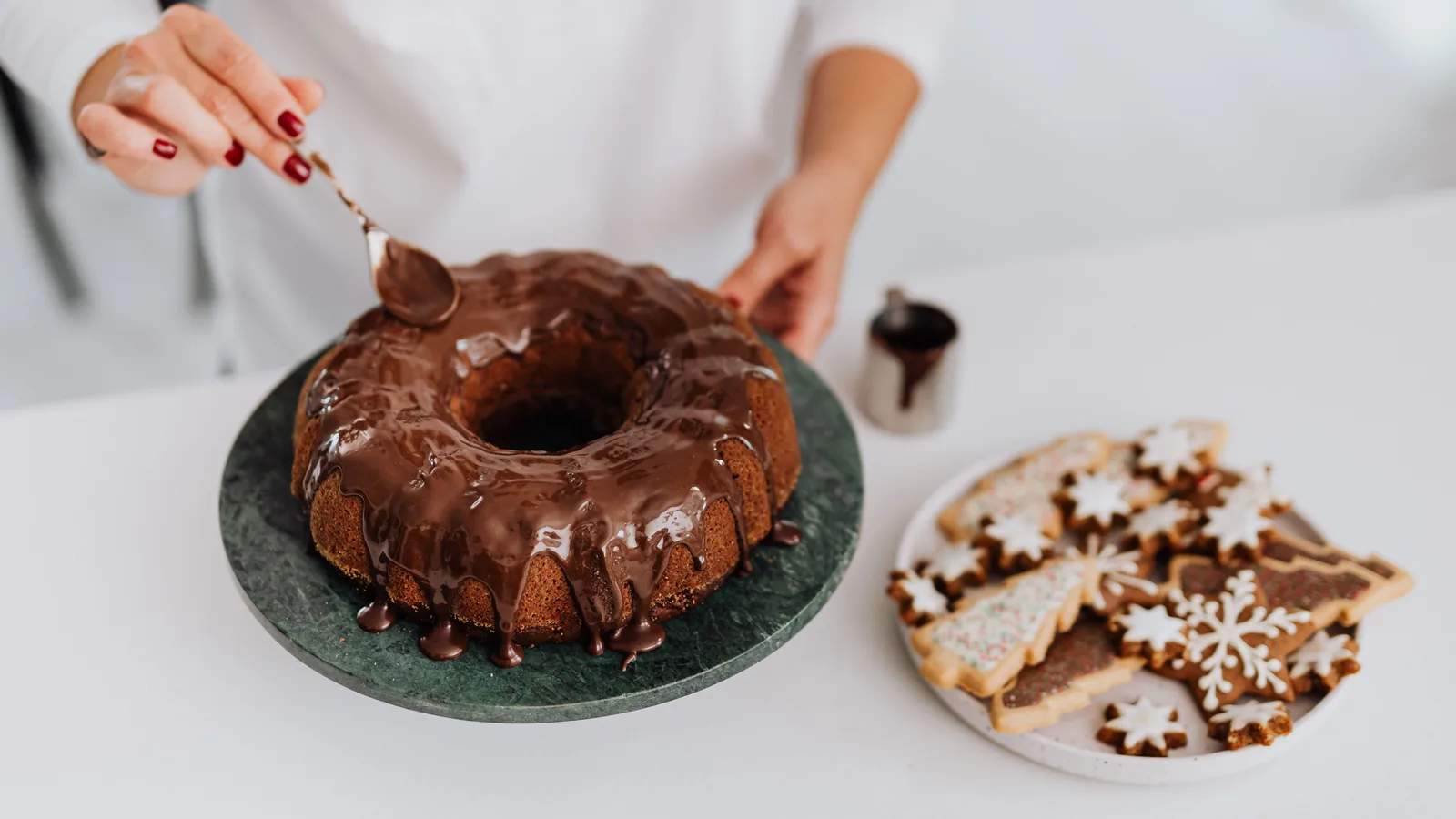 Uma mulher decora um bolo com cobertura de chocolate ao lado de biscoitos natalinos festivos.